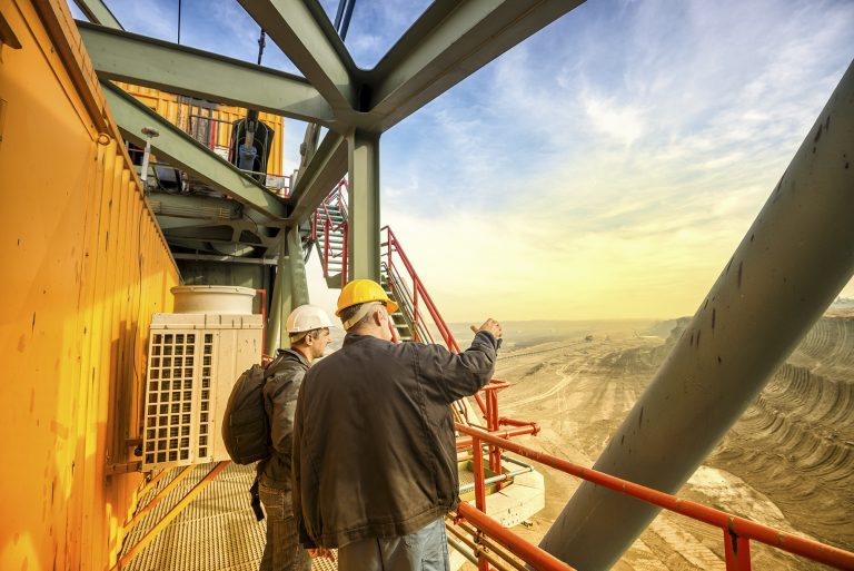 Two coal mine engineers with protective helmets standing on a huge drill machine, chatting and watching at the digging site. Beautiful and colorful sky in the background. Rear view.