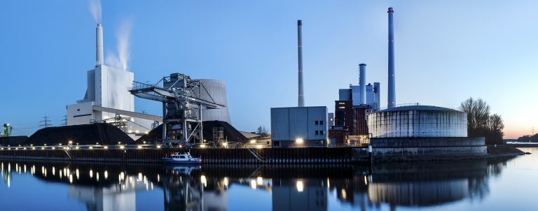 View of a coal-fired power plant in the early evening.  Water is seen in the foreground, showing the reflection of various features of the power plant.  There are numerous tall stacks seen on the property, and smoke is seen coming out of the ones to the left.  A blue sky is seen in the background.